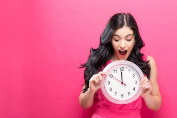 Junge Frau vor pinkem Hintergrund zeigt auf eine Uhr, wo es 5 vor 12 ist.