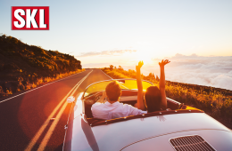 Lotterie-Gewinne im September 2020. Paar sitzt in einem silbernen Cabrio und fährt auf einer weiten Straße dem Sonnenuntergang entgegen.